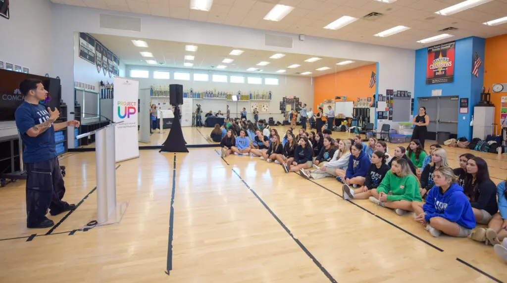 Students sitting on the floor listen to a man give a speech