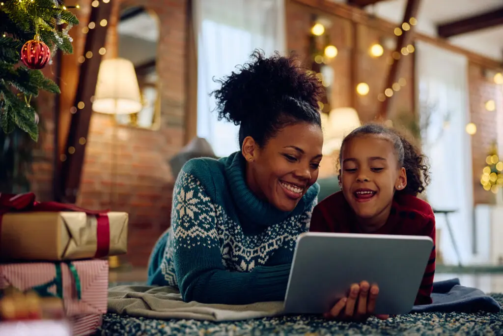 Family relaxing under holiday lights, browsing Black Friday and Cyber Monday deals on a laptop for holiday savings.