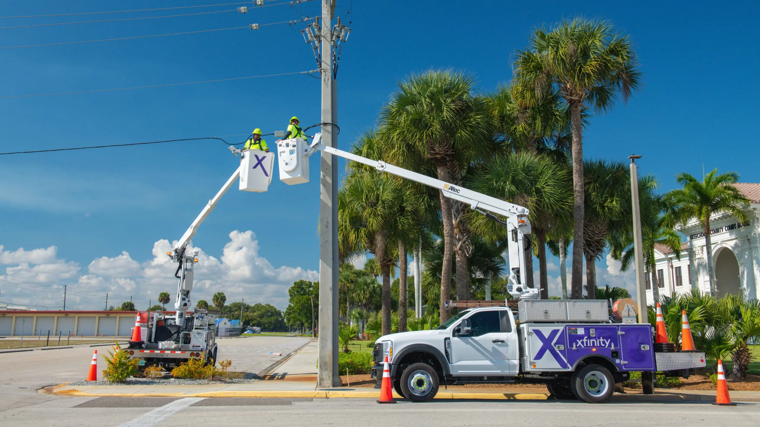 Two utility technicians wearing high-visibility safety gear install a network component on overhead cables using bucket trucks under a clear blue sky.
