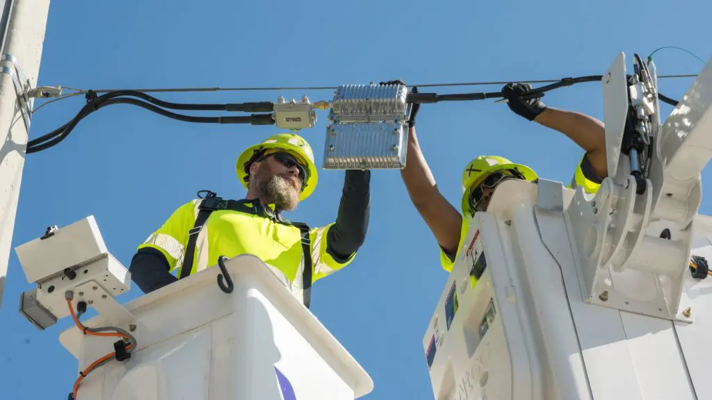 Two utility technicians wearing high-visibility safety gear install a network component on overhead cables using bucket trucks under a clear blue sky. Xfinity bucket trucks installing overhead network equipment on utility poles in Florida to expand Comcast’s high-speed Internet service.