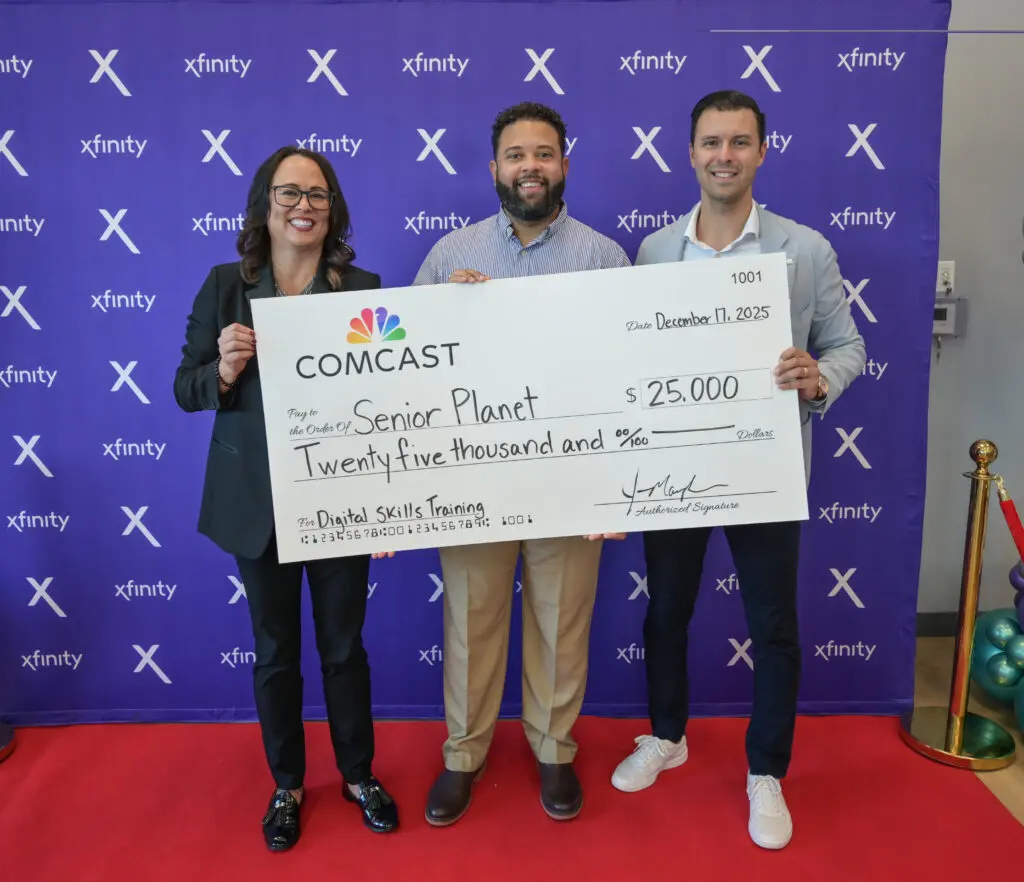 Three individuals holding an oversized check for $25,000 from Comcast to Senior Planet, standing in front of a purple Xfinity backdrop.