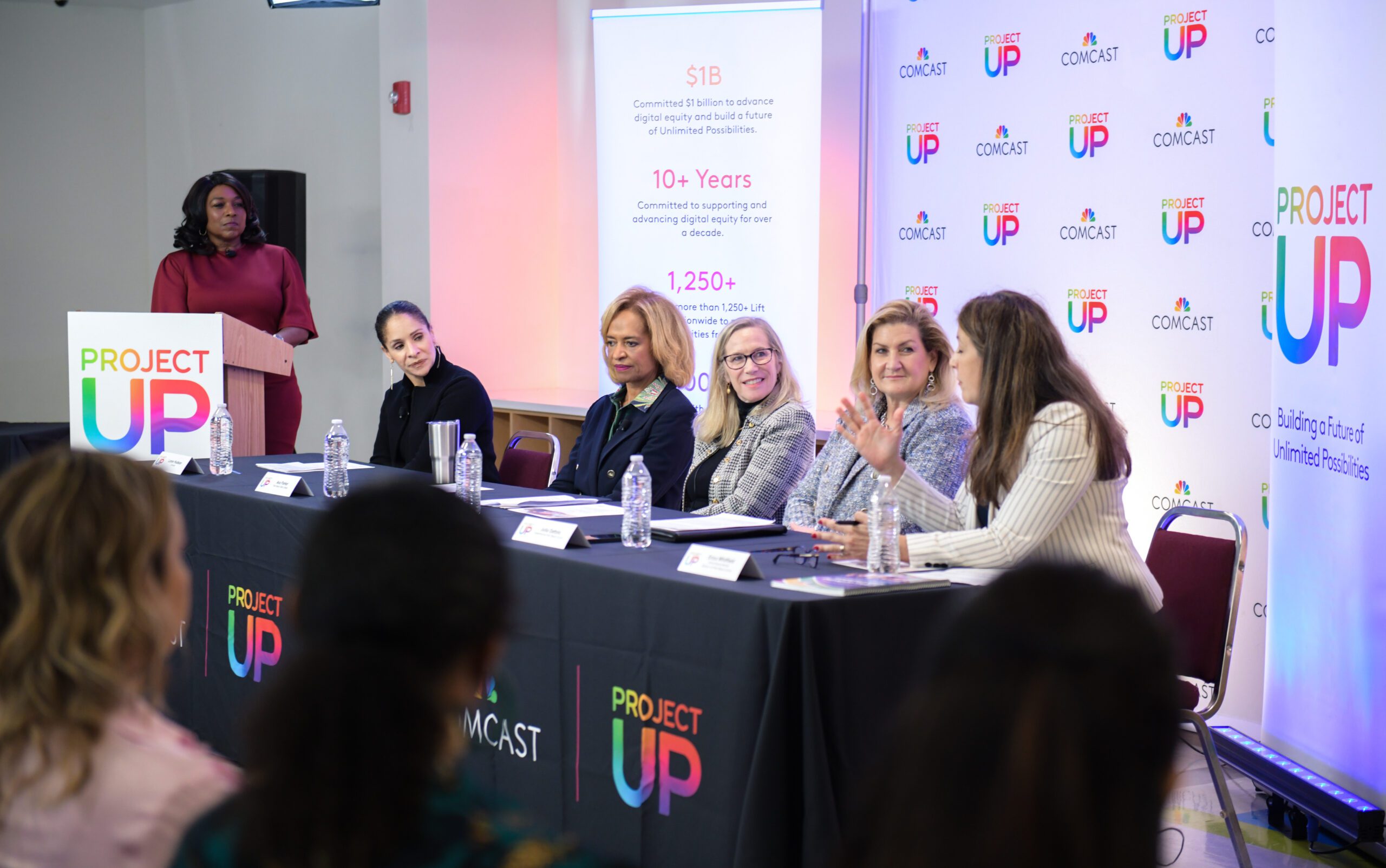 Women on panel are seated at a table. Moderator looks on from a podium.