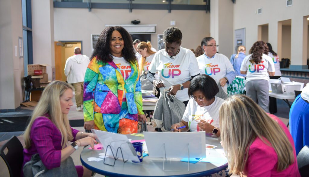 A group of Team UP volunteers sit at a table and write notes. One is standing and smiling at the camera.