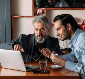 two men are sitting at a table, working on a laptop