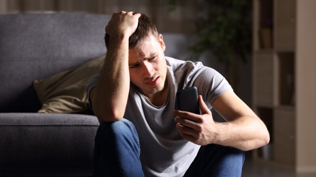 Single sad man checking mobile phone sitting on the floor in the living room at home with a dark background