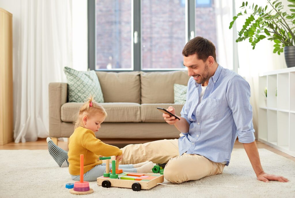 Xfinity Mobile customer in Florida enjoys fast WiFi on their device on the Premium Unlimited plan. IMAGE: Happy father with smartphone and little baby daughter playing with wooden toy toy blocks kit at home