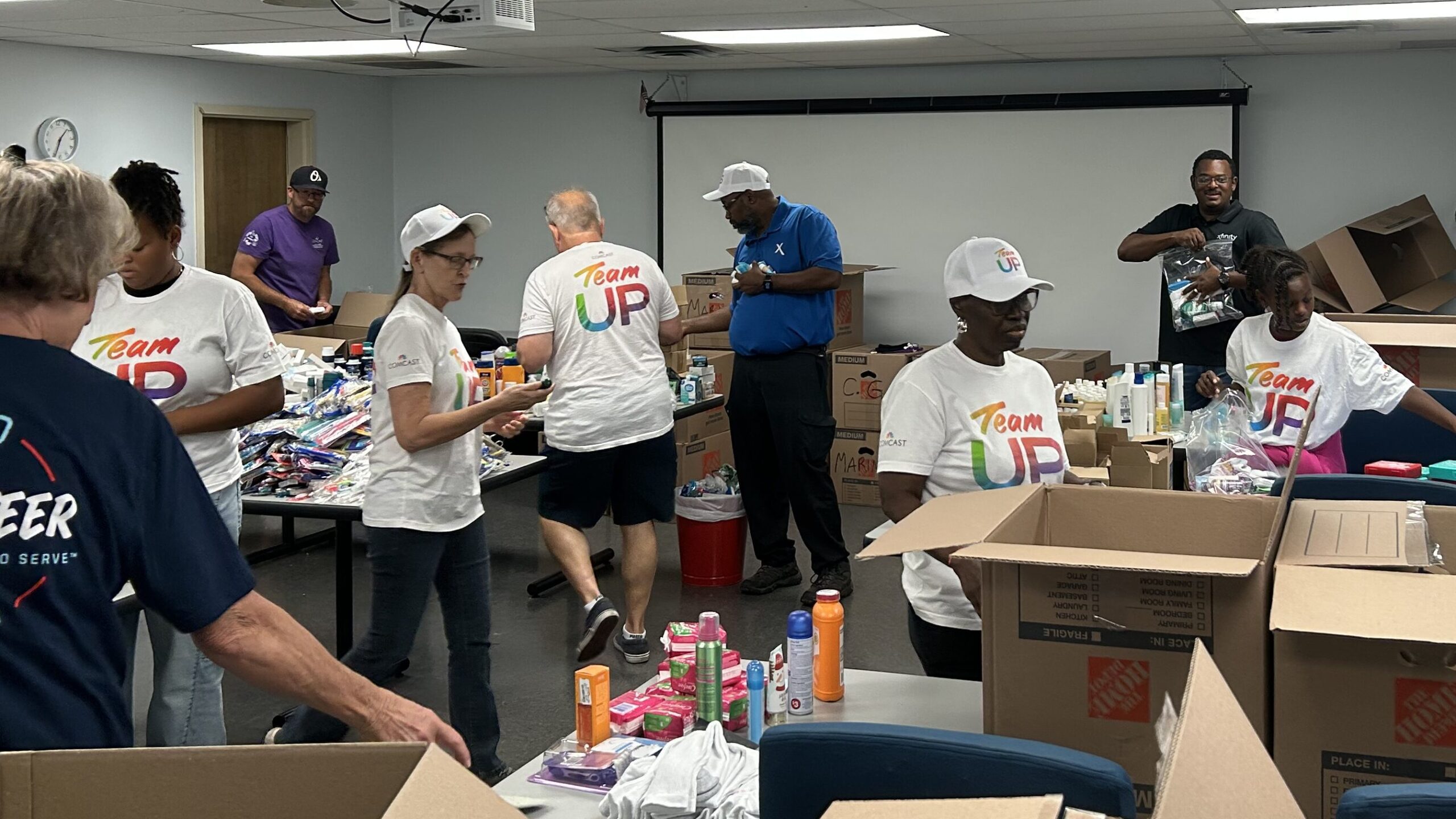 Comcast volunteers in Team UP shirts actively packing hygiene kits at tables inside the USO Jacksonville center, with boxes and supplies visible around them.