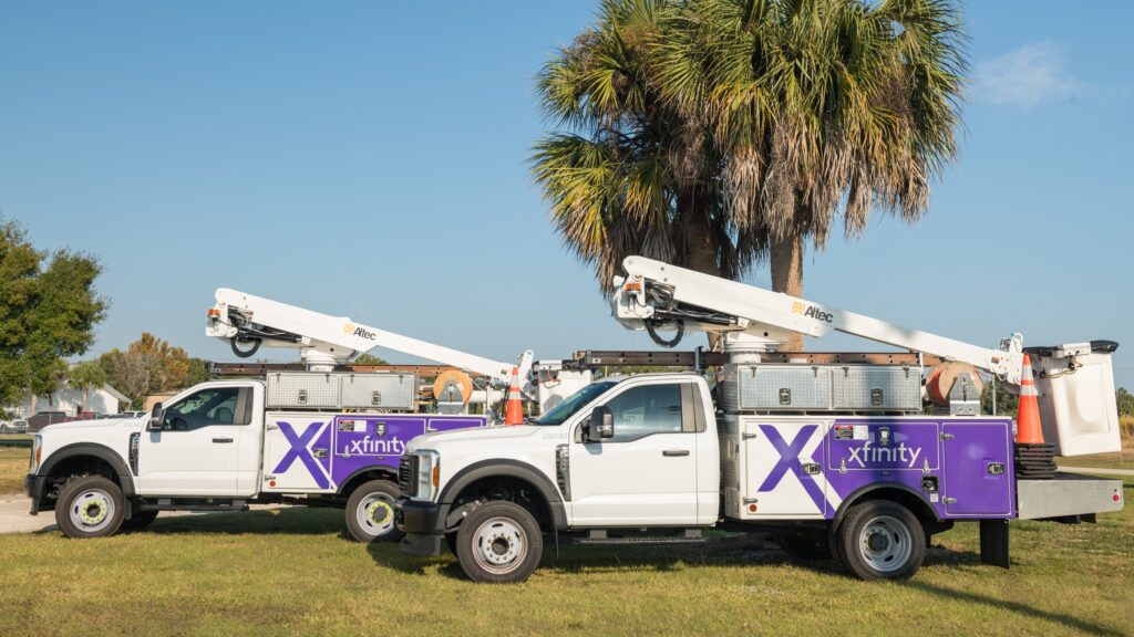 Two Xfinity-branded utility trucks with mounted bucket lifts parked on grass near palm trees, prepared for network maintenance or installation. Two Xfinity service trucks parked near palm trees in Florida, ready for network maintenance and Internet expansion projects.