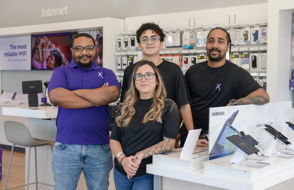 Four individuals standing inside the Xfinity store near a display of Samsung devices, with shelves of accessories in the background.