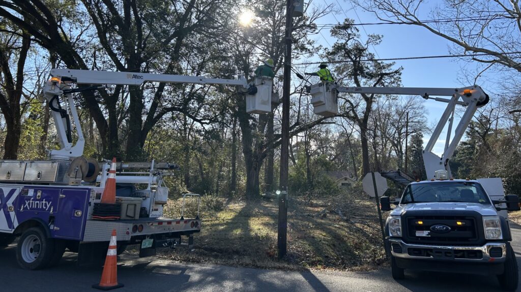 Two technicians in bucket trucks on opposite sides of a utility pole perform repairs on overhead lines, with service vehicles parked along a wooded roadside.