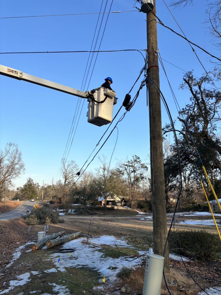 A technician in a bucket truck works on overhead utility lines beside a wooden pole in a rural area with patches of snow on the ground.